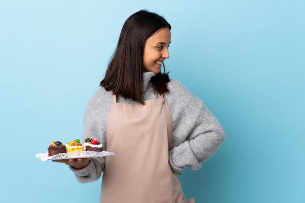 Foto chef pastelero sosteniendo un gran pastel sobre la pared azul mirando hacia un lado y sonriendo.