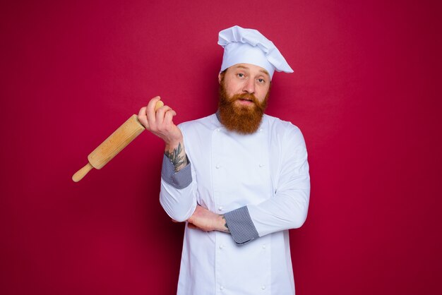 Chef pensativo con barba y delantal rojo chef sostiene un rodillo de madera