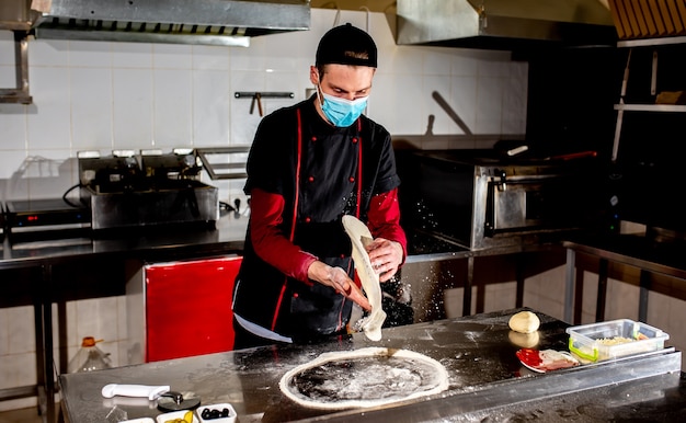El chef de pizza o el fabricante de una máscara médica contra el coronavirus, hace espacios en blanco de masa de pizza. Concepto de fabricación de masa de pizza