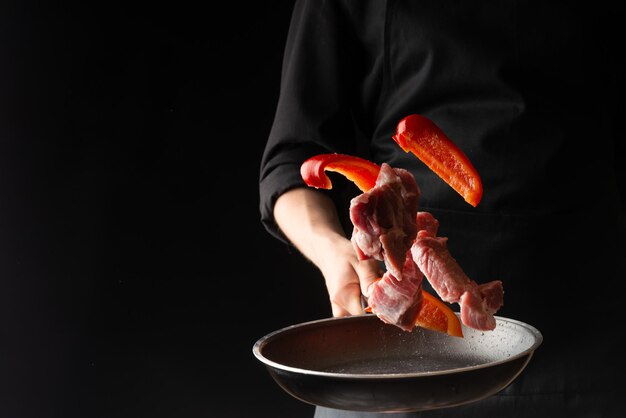 Foto el chef prepara bistecs de carne de cerdo o ternera arrojando carne fresca con pimientos dulces en una plancha sobre un fondo negro menú de fotos horizontal libro de recetas restaurantes comida sabrosa