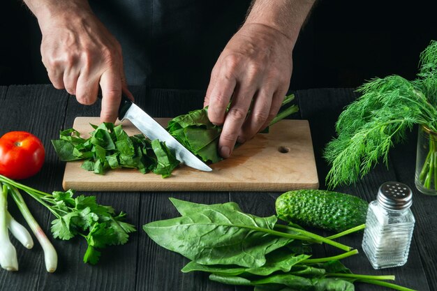 Un chef prepara una ensalada de espinacas en la cocina de un restaurante. Primer plano de las manos de un cocinero cortando verduras con un cuchillo sobre una tabla de cortar.