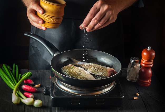 El chef prepara pescado fresco en una sartén espolvoreando sal con los ingredientes. Preparación para cocinar comida para peces. Entorno de trabajo en la cocina del restaurante.