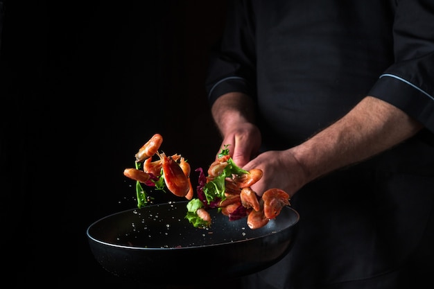 Un chef profesional cocina camarones en una sartén con verduras. Cocinar mariscos, comida vegetariana saludable y comida sobre un fondo oscuro. Congelamiento en movimiento. Espacio publicitario gratuito