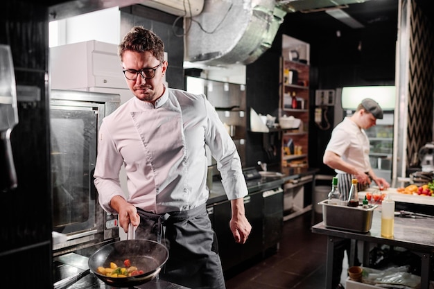 Chef profesional en delantal usando cocina para freír verduras en una sartén durante el trabajo en la cocina con un colega en segundo plano.