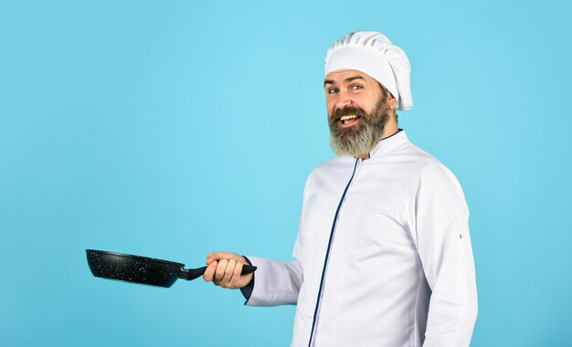 Chef profesional esposo con sombrero de chef en la cocina Concepto de comida de cocina Sostenga la sartén Cocinero barbudo de la mejor calidad en uniforme con herramientas Hombre barbudo Sostenga la sartén preparando la comida en la cocina