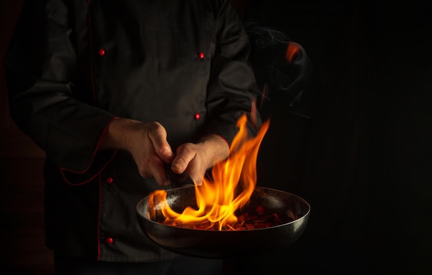 Chef profesional prepara comida vegetal en una sartén con llama de fuego