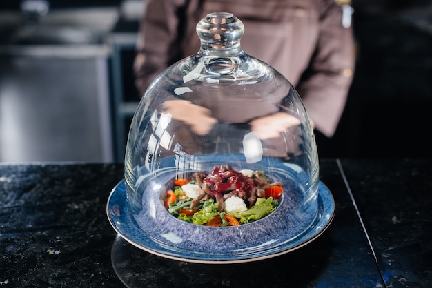 Un chef profesional sirve una ensalada recién preparada de tomate y verduras de ternera con salsa en un refinado restaurante bajo una cúpula de cristal.