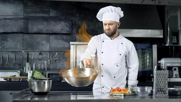 Chef profesional tirando comida en una sartén con llamas ardientes en la cocina