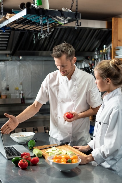 Chef profissional com tomate fresco explicando à sua estagiária o que fazer depois de picar vegetais, enquanto os dois estão de pé perto da mesa