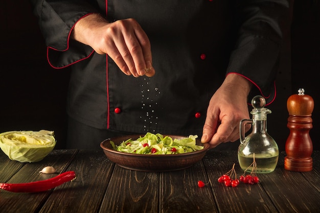 Chef profissional polvilha sal para salada de legumes frescos na mesa de madeira