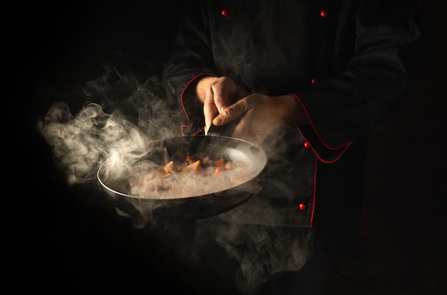 Chef profissional prepara comida em uma frigideira com vapor em um fundo preto