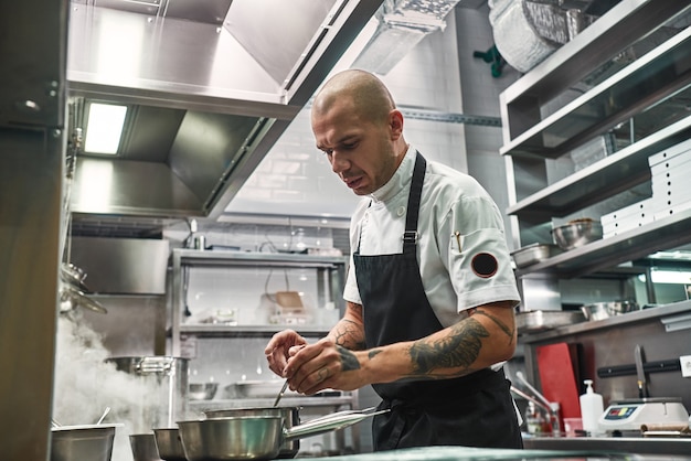 Chef profissional sério de avental com várias tatuagens nas mãos, cozinhando na cozinha de um restaurante