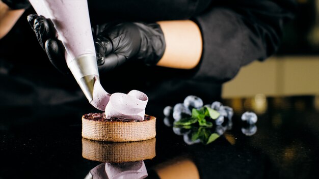 El chef de repostería decora la galleta con crema púrpura de la manga pastelera, primer plano.