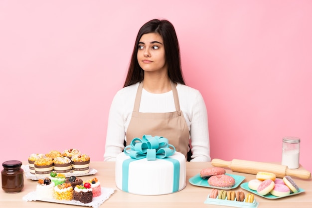 Chef de repostería con un gran pastel en una mesa sobre rosa aislado haciendo gesto de dudas mirando de lado