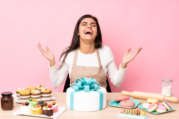 Chef de repostería con un gran pastel en una mesa sobre rosa aislado sonriendo mucho