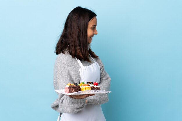 Chef de repostería sosteniendo un gran pastel sobre fondo azul aislado en posición lateral.