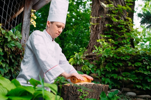 Un chef de restaurante en uniforme extiende masa sobre un tocón en el bosque