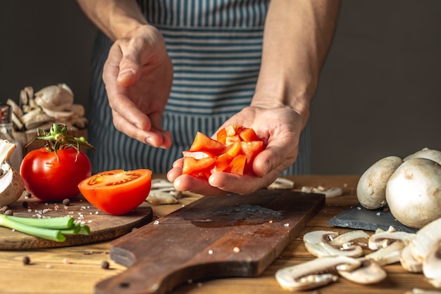 Un chef de sexo masculino con un delantal azul está cortando apetitosos tomates frescos con un cuchillo para preparar un plato. Concepto del proceso de cocción.