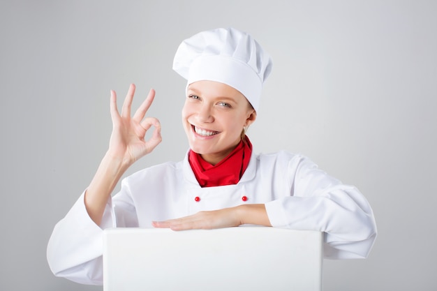 Chef Sign. Cocinero / panadero de la mujer que mira sobre la cartelera de papel de la muestra. Mujer de expresión sorprendida y divertida aislada en la pared blanca.