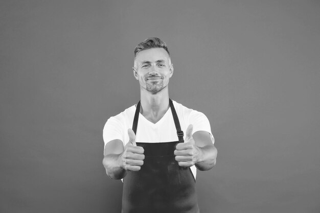 El chef sonriente muestra el pulgar hacia arriba. Chef profesional busca chef masculino en delantal rojo. El hombre de negocios usa delantal en la cafetería. Su trabajo favorito.