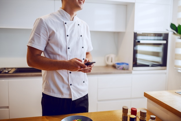 Chef sonriente en uniforme blanco de pie en la cocina junto a la encimera y usando el teléfono inteligente.