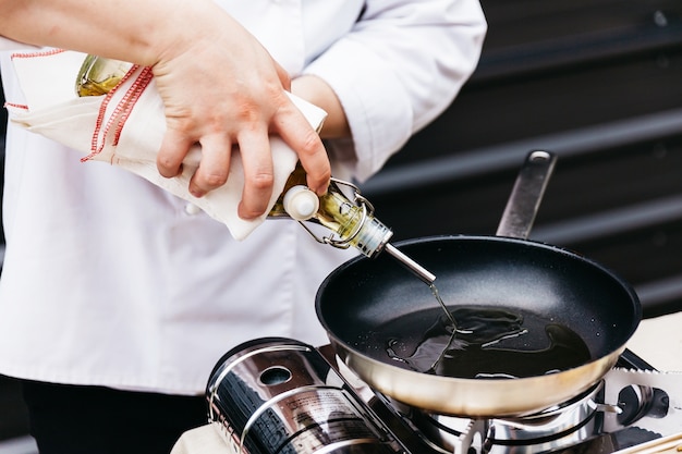 Chef sosteniendo una botella de aceite de oliva que envuelve con tela mientras vierte aceite de oliva en una sartén caliente.