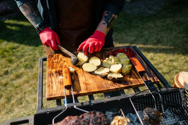 Chef tatuado con guantes de goma cortando papas a la parrilla al aire libre Concepto de fiesta a la parrilla