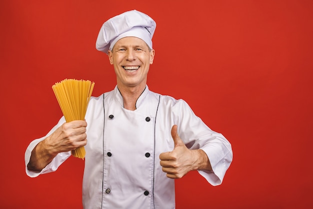 Chef tiene manojo de espagueti en la mano. Concepto de catering y comida italiana aislado sobre fondo rojo. Cocinar con la cara satisfecha en uniforme blanco tiene pasta seca mostrando los pulgares hacia arriba.