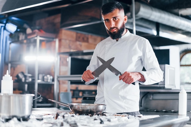 Foto chef en uniforme blanco de pie en la cocina sosteniendo cuchillos en las manos