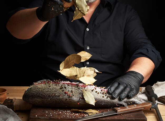 Chef en uniforme negro y guantes de látex negro vierte hojas secas de laurel sobre filete de salmón fresco cortado