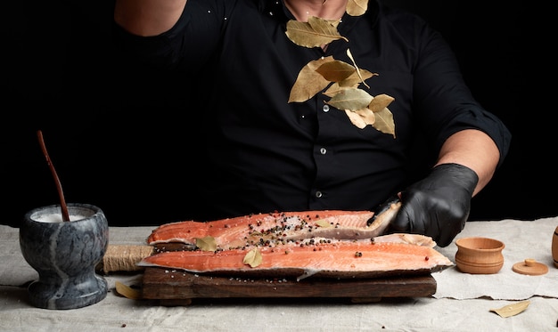 Chef en uniforme negro y guantes de látex negro vierte hojas secas de laurel sobre filete de salmón fresco cortado