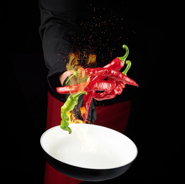 Foto chef en uniforme negro sostiene una sartén redonda y arroja chiles enteros rojos y verdes en un fuego ardiente