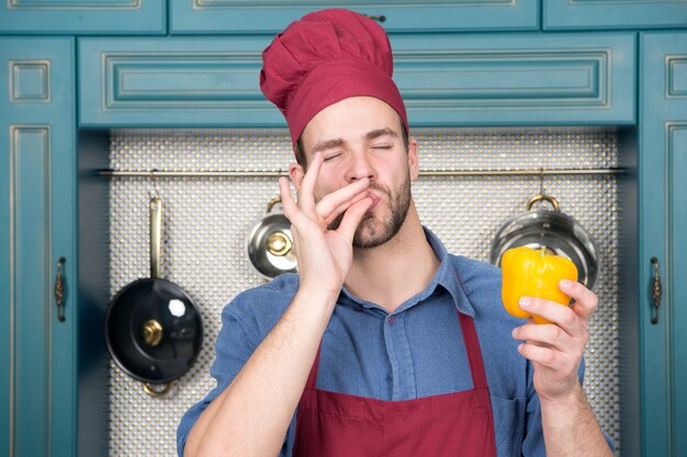 Chef zeigt ok Geste zum Paprika. Kochmann mit rotem Hut, Schürze schmeckt Gemüse in der Küche. Vegetarisch, Gesundheit, Ernährung, Vitamin. Essen, Kochen, Küche, Menü, Gerichte, Rezepte.