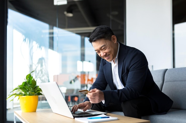 Chefe asiático transfere dinheiro de um cartão bancário sentado no escritório satisfeito com o resultado que o empresário sorri trabalha no laptop