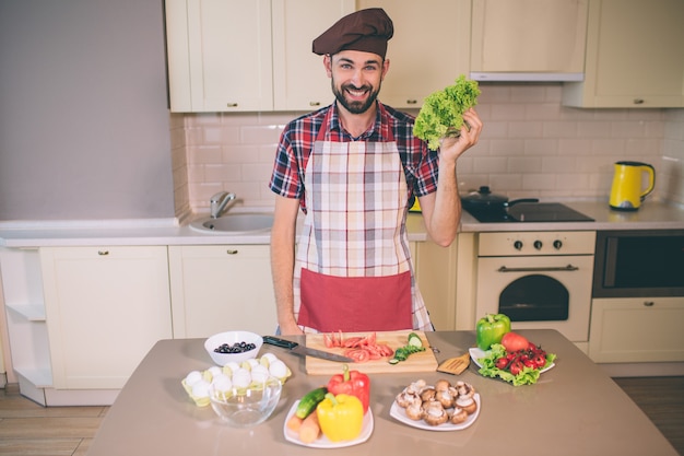 Chefe feliz e positivo fica na cozinha e parece. Ele sorri. Jovem mantém alface na mão. Ele usa pron. Há ovos e legumes na mesa.