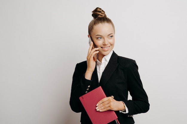 Chefin in eleganter klassischer Kleidung, die mit zufriedenem Blick richtig aussieht, telefoniert und sich freut, gute Nachrichten über Arbeitsergebnisse zu hören, mit Notebook in der Hand einzeln auf grauem Hintergrund posieren