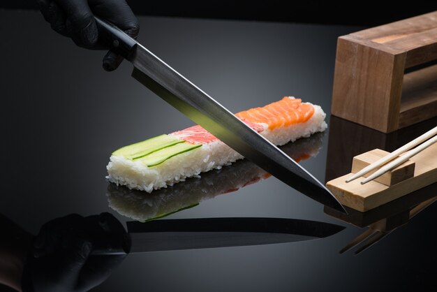 Chefkoch bereitet Sushi zu, schneidet mit einem Messer. Sushi auf schwarzem Hintergrund mit Reflexion