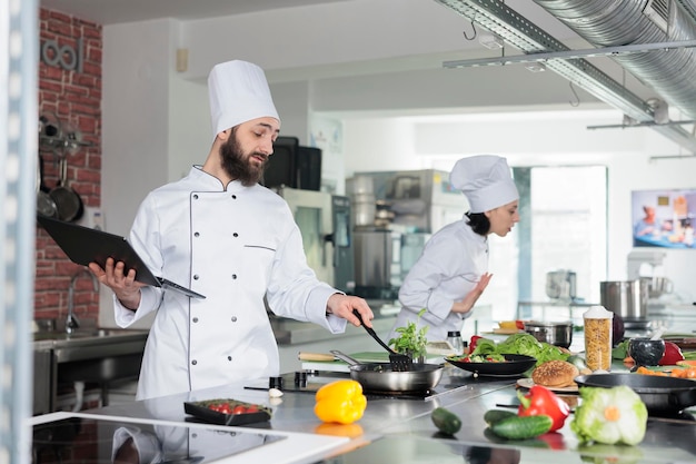 Chefkoch der Gourmetküche kocht Abendessen, während er den Computer verwendet, um das Rezept genau zu befolgen. Gastronomischer Experte bereitet Gerichte mit frischen Bio-Zutaten zu, während er einen Laptop in der Küche hält.