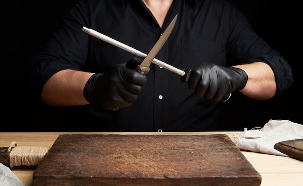 Chefkoch in einem schwarzen Hemd und schwarzen Latexhandschuhen schärft ein Küchenmesser auf einem Eisenschärfer