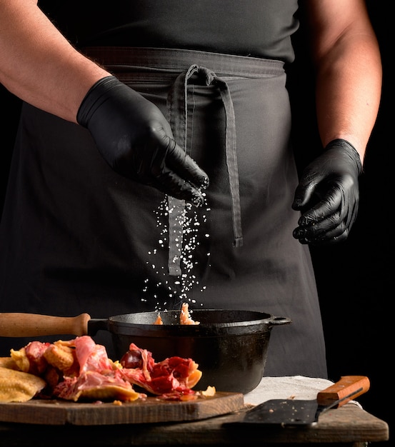 Chefkoch in schwarzer Uniform und mit Latexhandschuhen bestreut mit weißem Salz rohes Hühnerfleisch in einer schwarzen gusseisernen Pfanne