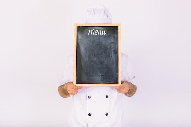 Chefkoch trägt Küchenjacke und Hut, der sein Gesicht mit einer Tafel mit der Speisekarte bedeckt, auf weißem Hintergrund.