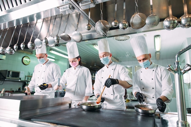 Chefs com máscaras protetoras e luvas preparam os alimentos na cozinha de um restaurante ou hotel.