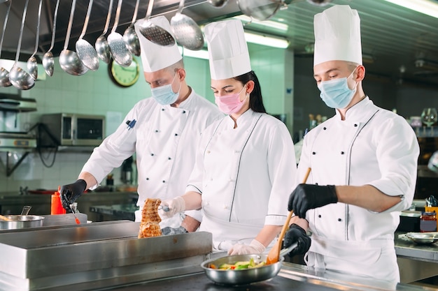 Chefs com máscaras protetoras e luvas preparam os alimentos na cozinha de um restaurante ou hotel.