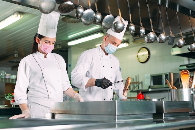 Los chefs con máscaras protectoras y guantes preparan la comida en la cocina de un restaurante u hotel.