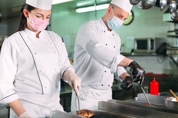 Los chefs con máscaras protectoras y guantes preparan la comida en la cocina de un restaurante u hotel.