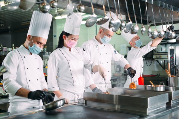 Los chefs con máscaras protectoras y guantes preparan la comida en la cocina de un restaurante u hotel.