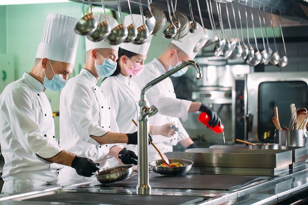 Los chefs con máscaras protectoras y guantes preparan la comida en la cocina de un restaurante u hotel.