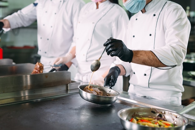 Chefs con máscaras protectoras y guantes preparan la comida en la cocina de un restaurante u hotel.