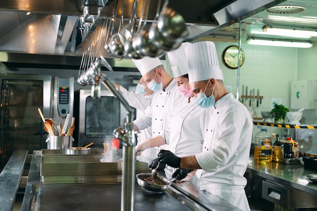 Chefs con máscaras protectoras y guantes preparan la comida en la cocina de un restaurante u hotel.