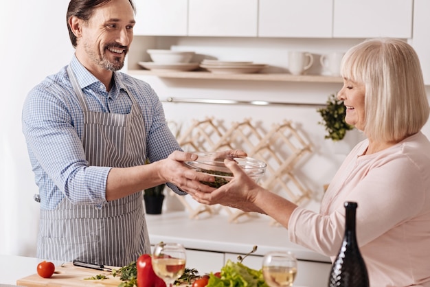 Cheio de positividade. Homem barbudo feliz positivo em pé na cozinha aproveitando o fim de semana enquanto cozinha uma salada com sua mãe sênior